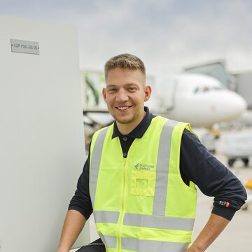 Ein junger Mann in dunkler Arbeitskleidung und neongelber Warnweste mit der Aufschrift „Stuttgart Airport“ steht draußen auf dem Flughafengelände. Er lächelt freundlich in die Kamera und lehnt mit einem Bein an einem Technikgehäuse. Im Hintergrund ist ein Flugzeug zu sehen.